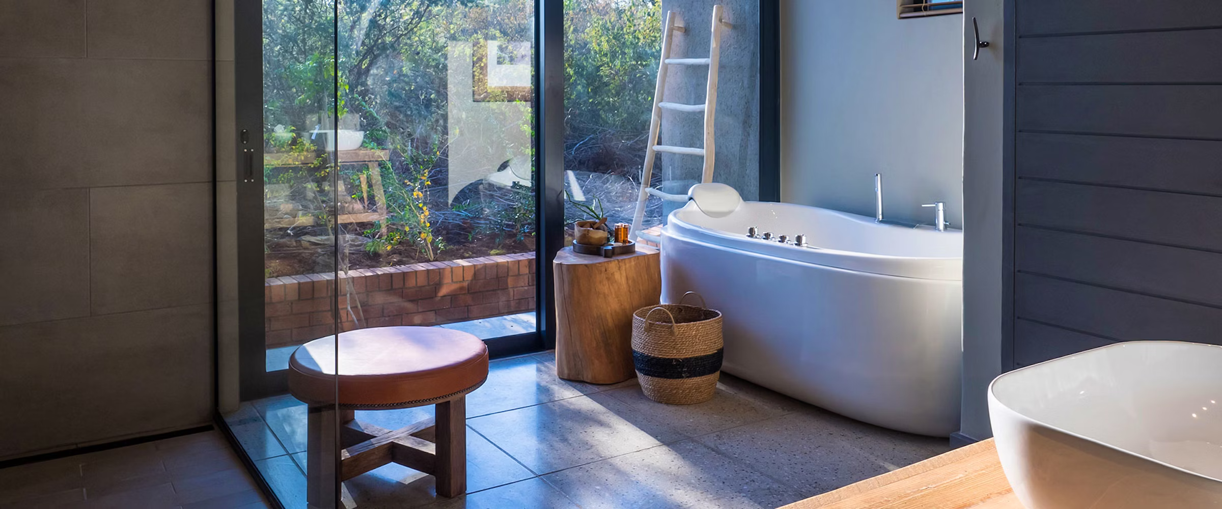 A modern bathroom featuring a sleek white bathtub, wooden stool, and natural accents. Large glass doors reveal a lush garden setting, enhancing the tranquil atmosphere. A woven basket and wooden table add warmth to the contemporary design.