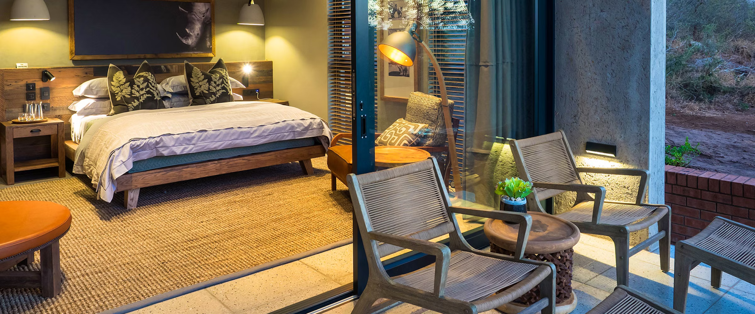 A warmly lit bedroom features a wooden bed with patterned pillows and bedside lamps. A large glass door opens to a patio with wicker chairs and a small table, overlooking a natural landscape. The room includes modern decor with a focus on comfort and nature.