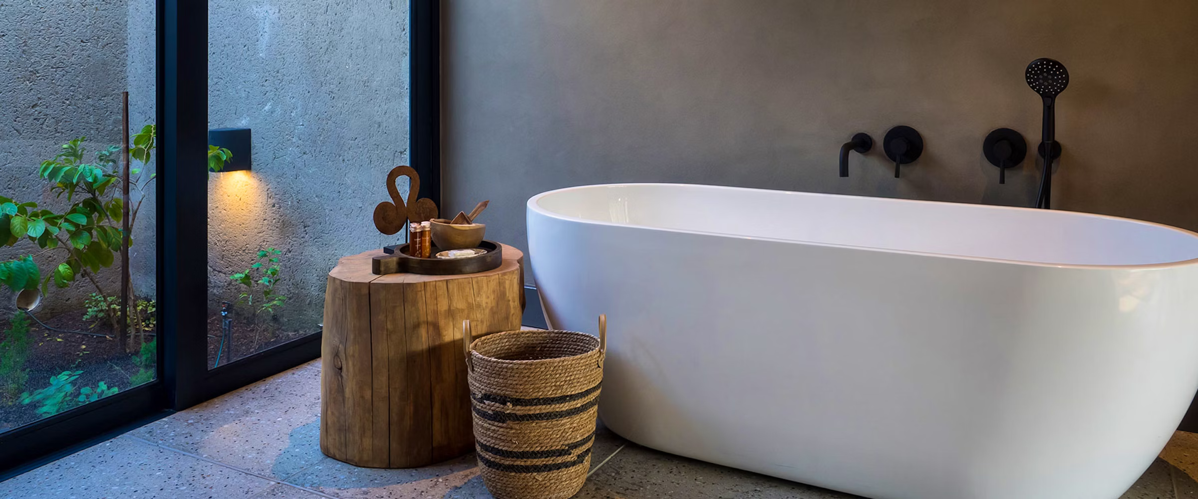 A sleek white bathtub in a minimalist bathroom with a large window showcasing a small garden. A rustic wooden stool holds bath items, and a woven basket sits nearby. The room features subtle lighting and a natural stone floor, creating a tranquil atmosphere.
