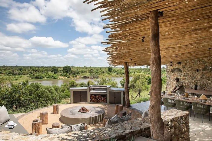 An outdoor lounge with a central fire pit surrounded by cushioned seating. The area features a rustic wooden roof supported by tree trunks, overlooking a lush landscape and river. A dining table is set on a stone patio under the roof, enhancing the natural ambiance.