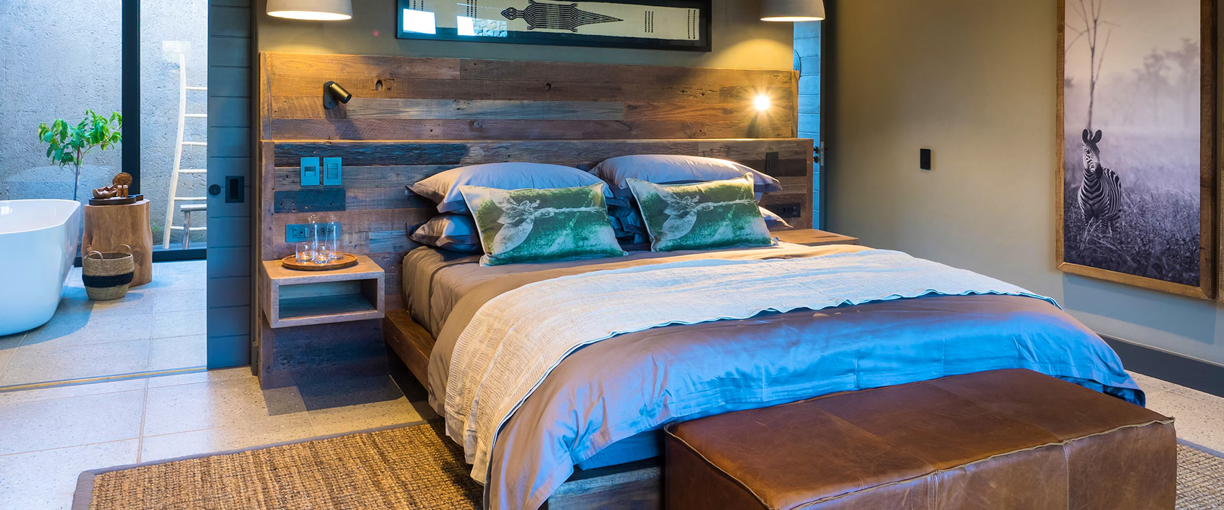 A cozy bedroom features a wooden headboard, plush bedding with green accent pillows, and a warm ambiance. Adjacent is a stylish bathroom with a freestanding tub and a potted plant. A zebra print adorns the wall, enhancing the room's natural charm.