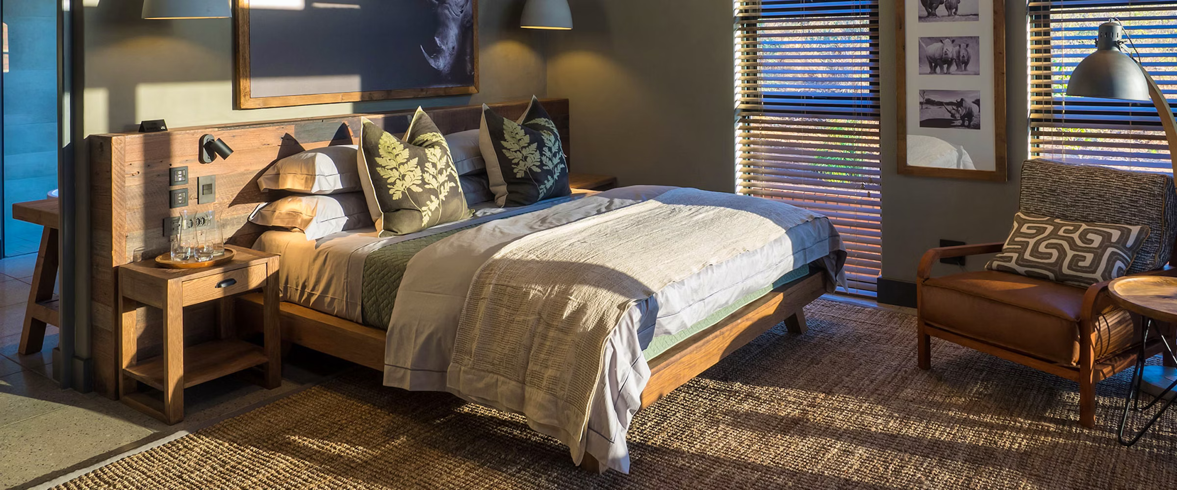 A cozy bedroom features a wooden bed with patterned pillows, a soft blanket, and a rustic nightstand holding water bottles. Sunlight streams through blinds, illuminating a cushioned armchair with a decorative pillow. Animal-themed artwork adorns the wall, enhancing the warm ambiance.