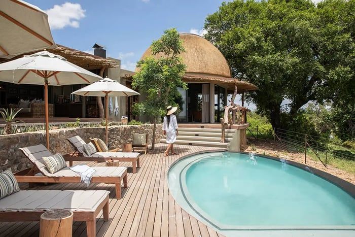 A woman in a sun hat walks by a kidney-shaped pool surrounded by wooden decking and lounge chairs with umbrellas. The setting is luxurious, with a thatched-roof building and lush greenery in the background, under a clear blue sky.