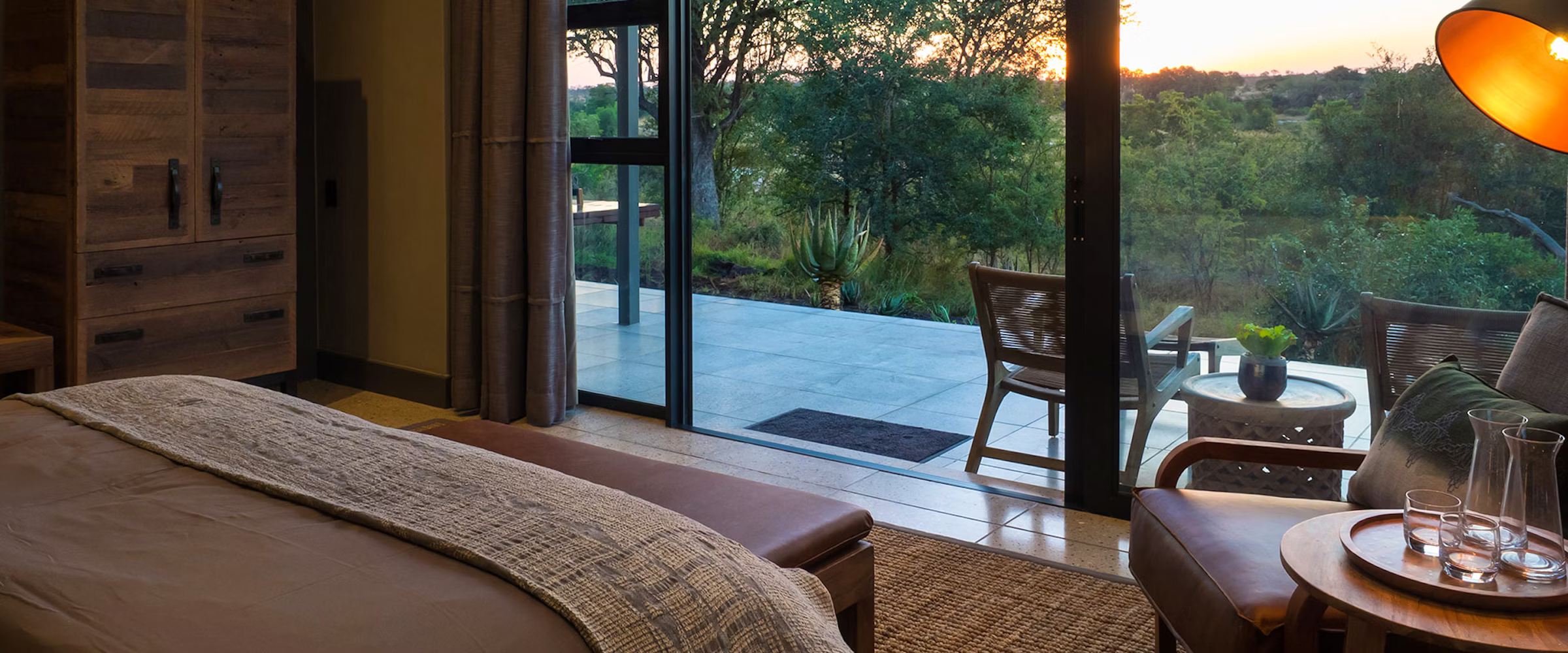 A tranquil bedroom featuring a large bed with a textured throw, a wooden wardrobe, and a stylish lamp. The room opens to a patio with seating and lush greenery visible through glass doors, offering a peaceful view of the garden at sunset.