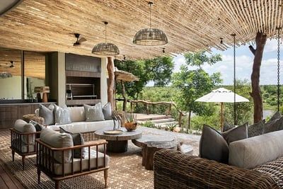 An elegant outdoor lounge featuring wicker furniture, a rustic wooden ceiling, and a view of lush greenery. The space includes comfortable sofas, a coffee table, and a barbecue setup, creating a relaxing environment under pendant lights and natural light filtering through the foliage.