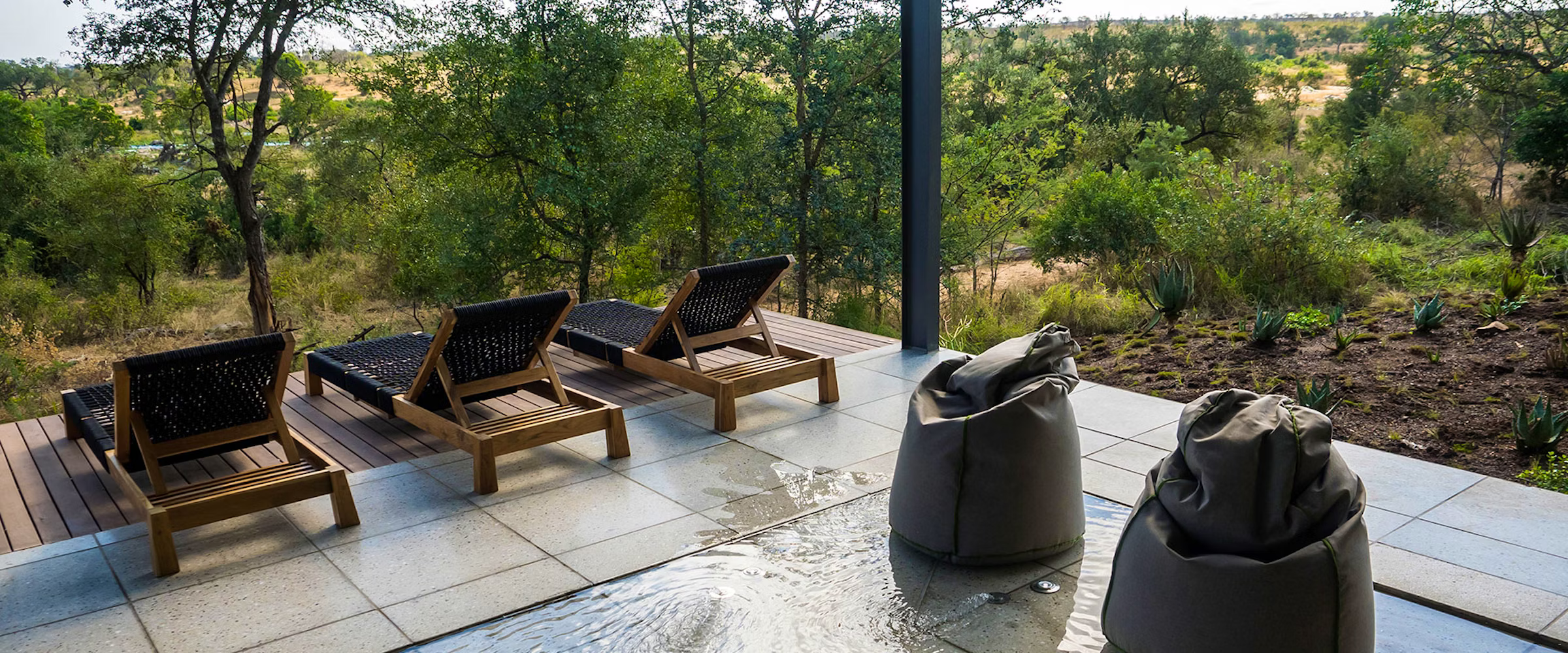 A tranquil terrace with three wooden lounge chairs and two bean bags, overlooking a lush green forest. The setting features a stone patio leading to a natural landscape, creating a peaceful outdoor retreat.
