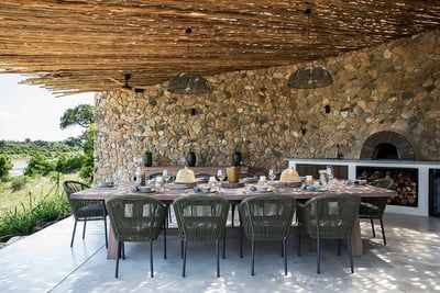 A rustic outdoor dining space featuring a long wooden table set with plates, glasses, and decorative items. The area is covered by a wooden pergola and backed by a stone wall with a built-in wood-fired oven. Lush greenery and a river are visible in the background.