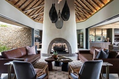 A modern rustic living room featuring a central round stone fireplace, leather sofas, and armchairs. The space has a high ceiling with exposed wooden beams and large windows, offering an open, airy feel. Decorative hanging lights and a patterned rug complete the cozy, stylish look.