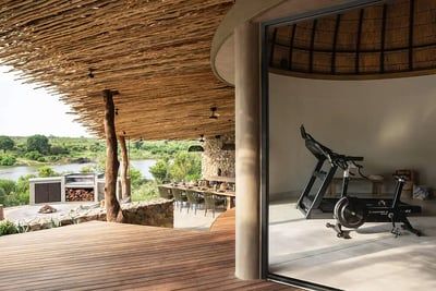 A modern gym with exercise equipment overlooks a serene landscape with lush greenery and a river. The gym features a thatched roof and is part of a luxurious open-air building with wooden decking and a large dining table visible outside.