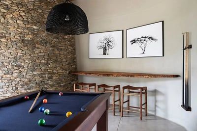 A chic pool room featuring a black pool table with colorful balls and cue sticks. The room has a textured stone wall, a woven black pendant light, and two framed tree art pieces. A wooden shelf with stools provides seating, and a cue rack is mounted on the wall.