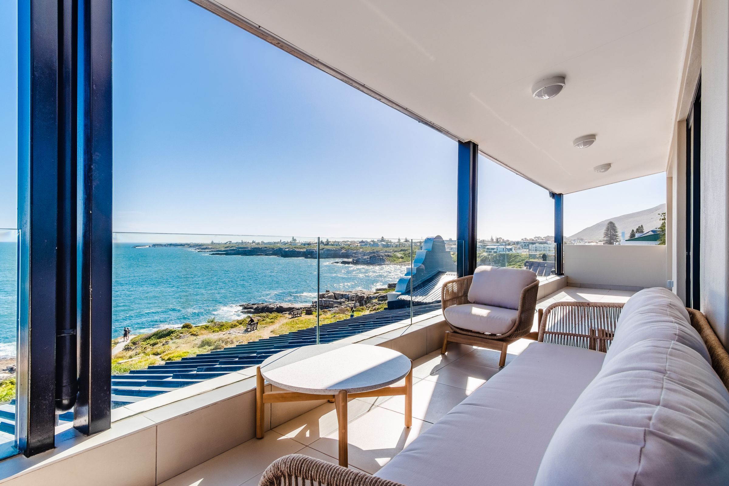 A spacious balcony with comfortable seating overlooks a stunning ocean view. The clear glass railing provides an unobstructed vista of the coastline and distant townscape, creating a serene and inviting outdoor space.