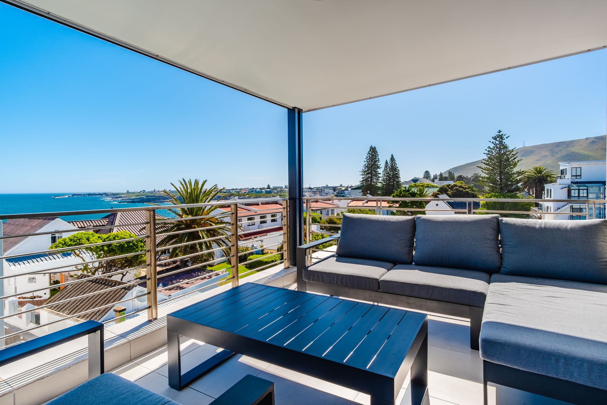 A spacious balcony features a modern outdoor lounge set with grey cushions and a sleek black coffee table. The area offers stunning views of the ocean and surrounding landscape, including greenery and rooftops, under a clear blue sky.