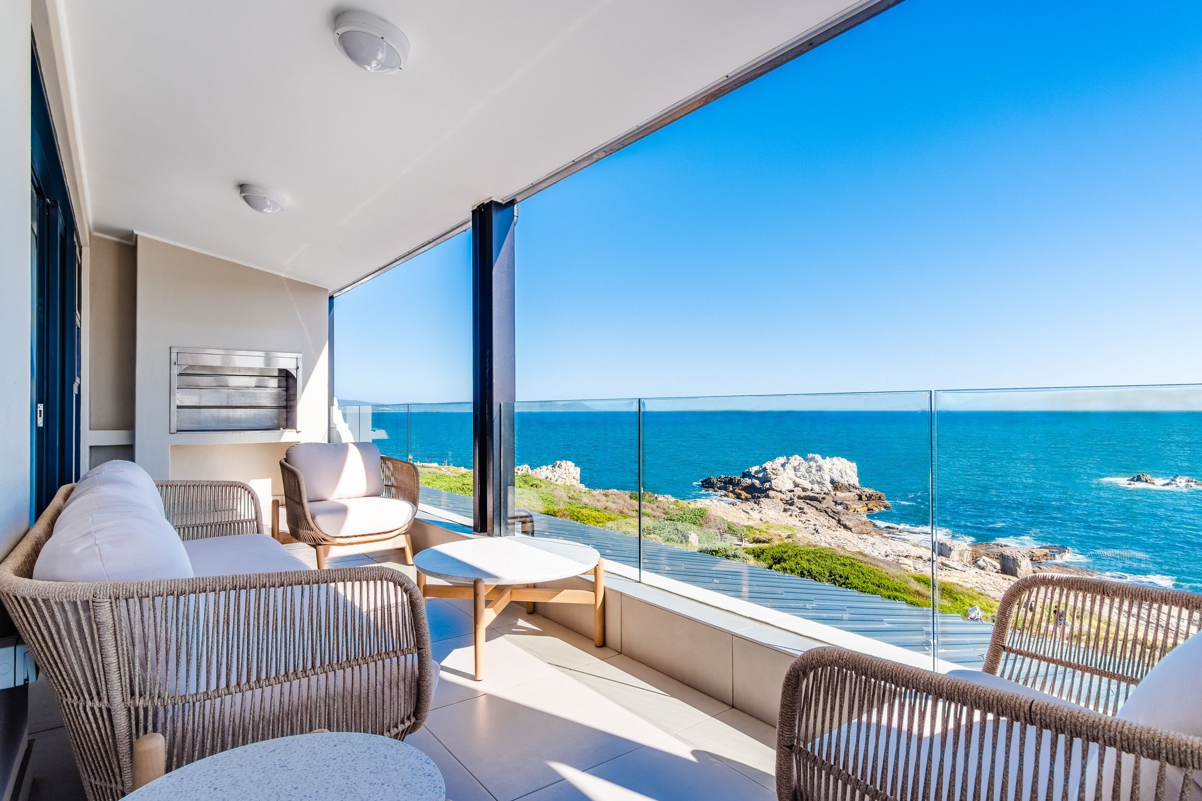 A spacious terrace with wicker seating and a glass railing offers stunning views of the ocean and rocky coastline under a clear blue sky, perfect for relaxation and enjoying the coastal scenery.