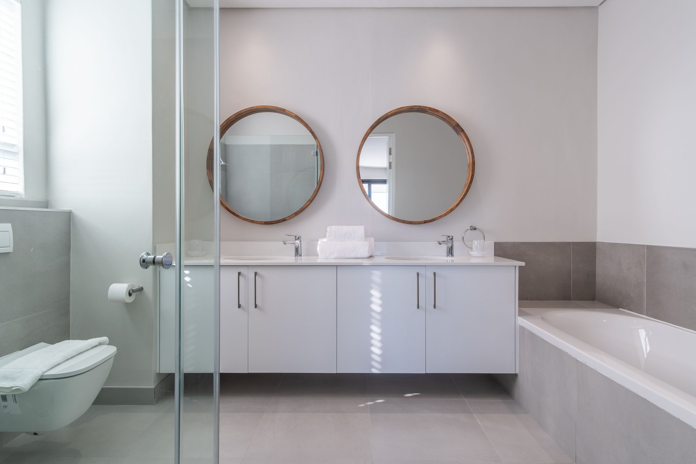 A spacious bathroom featuring dual round mirrors above a double-sink vanity with sleek faucets. The room includes a glass-enclosed shower and a built-in bathtub, complemented by modern gray tiling and natural light from a window.