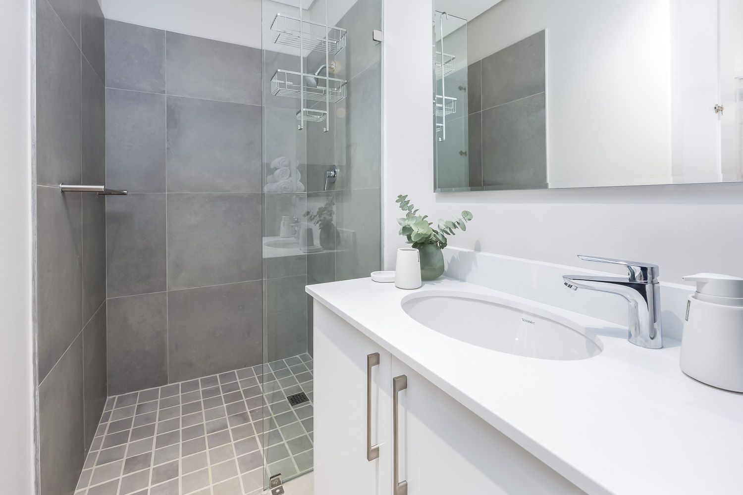 A contemporary bathroom featuring a spacious walk-in shower with gray tiles, a sleek white countertop with a built-in sink, and chrome fixtures. A small plant decorates the counter, adding a touch of greenery to the clean, minimalist space.