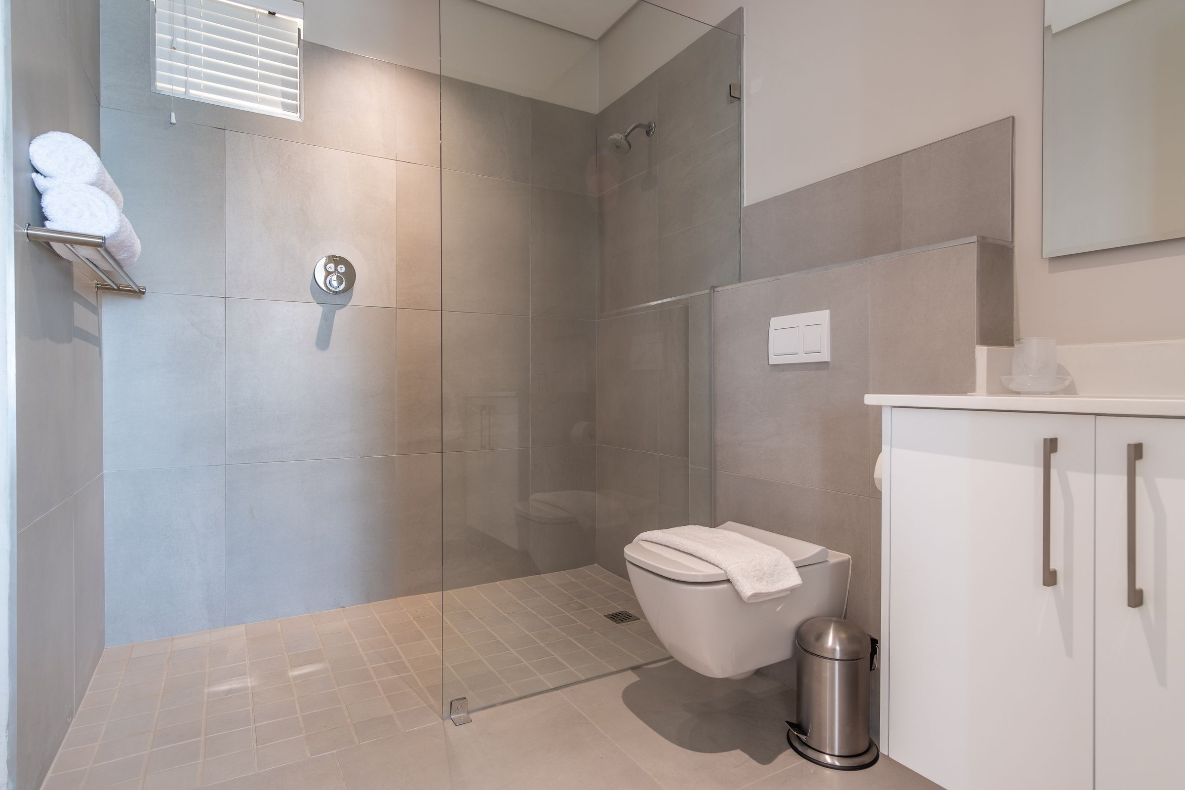 A sleek bathroom featuring a glass-enclosed shower, light gray tiles, a modern wall-mounted toilet, and a white countertop cabinet. A towel rack with folded towels and a small window allow natural light to enhance the minimalist decor.