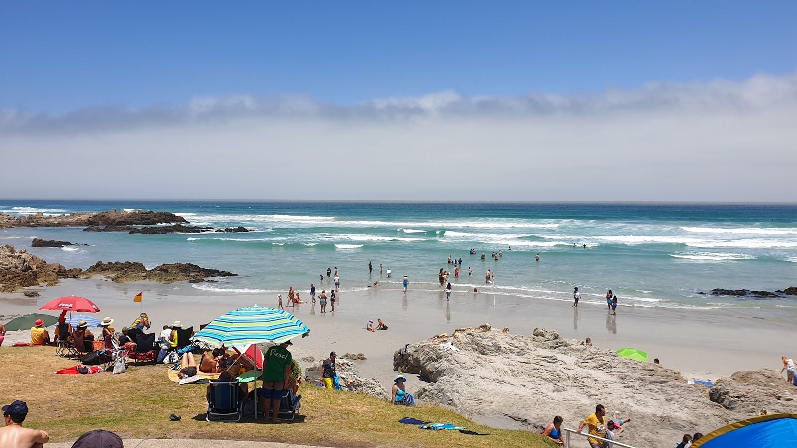 A vibrant beach scene with many people enjoying the sunny weather. The foreground shows groups under colorful umbrellas on the grassy area, while others swim and play in the ocean. Rocky outcrops are visible, and the sky is clear with a few clouds.