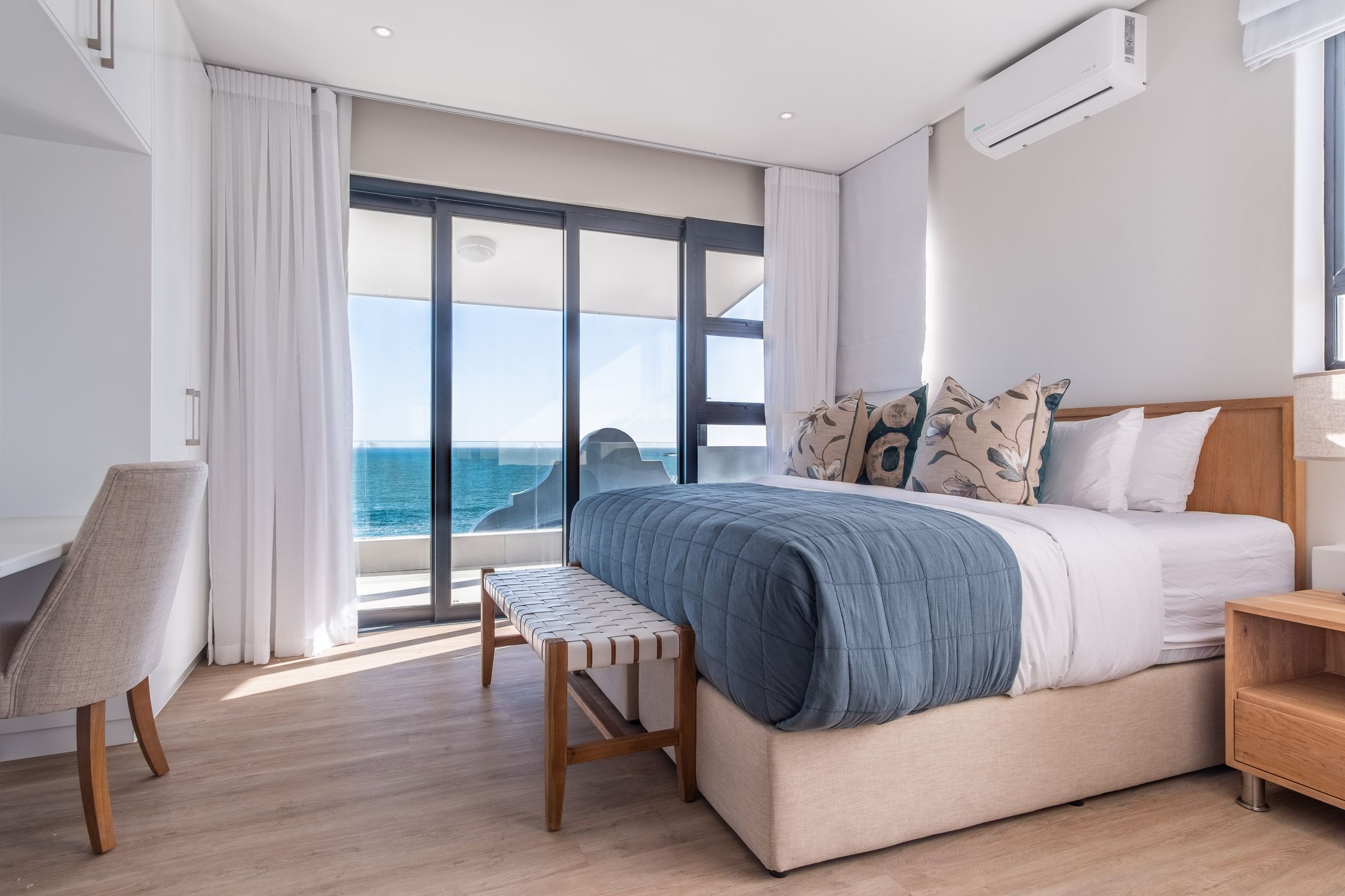 A serene bedroom with a large bed adorned with decorative pillows and a blue quilt. Floor-to-ceiling windows offer stunning ocean views. The room features light wood flooring, a small desk with a chair, and an air conditioning unit, creating a tranquil coastal ambiance.