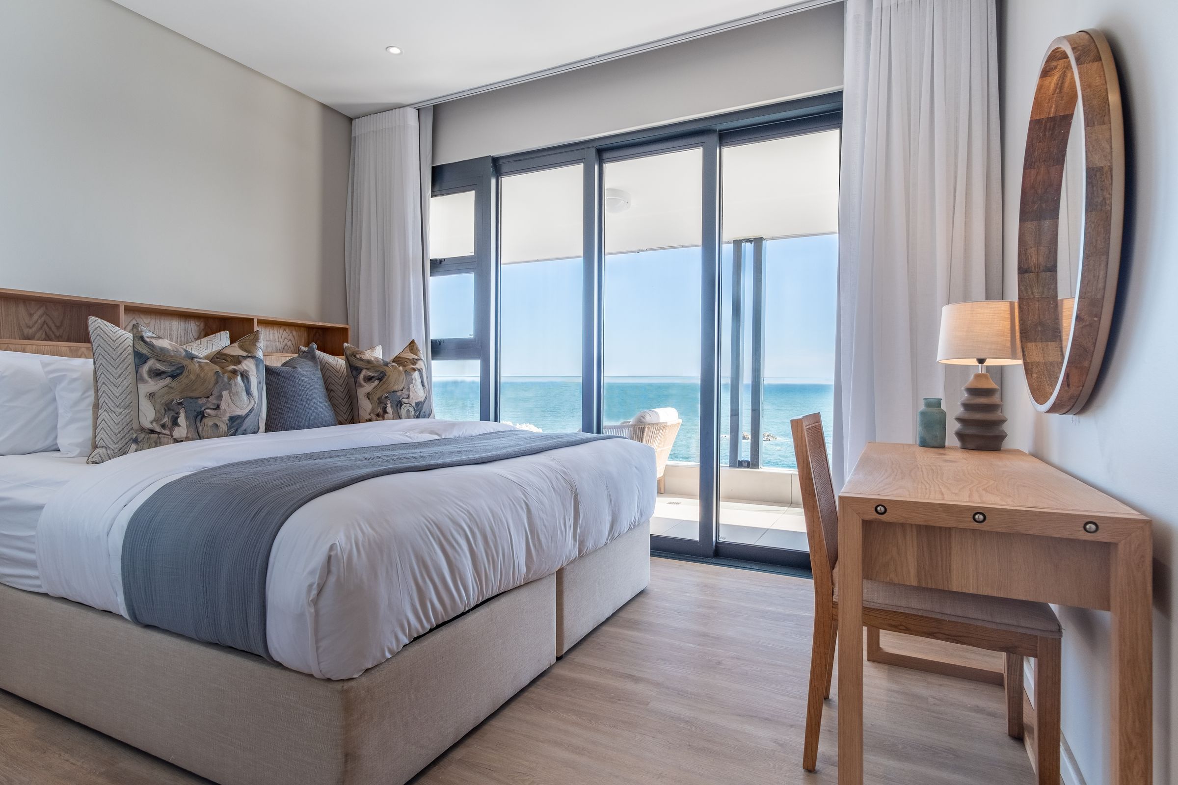 A stylish bedroom featuring a large bed with decorative pillows, a wooden desk with a mirror and lamp, and a stunning ocean view through floor-to-ceiling glass doors, complemented by light curtains.