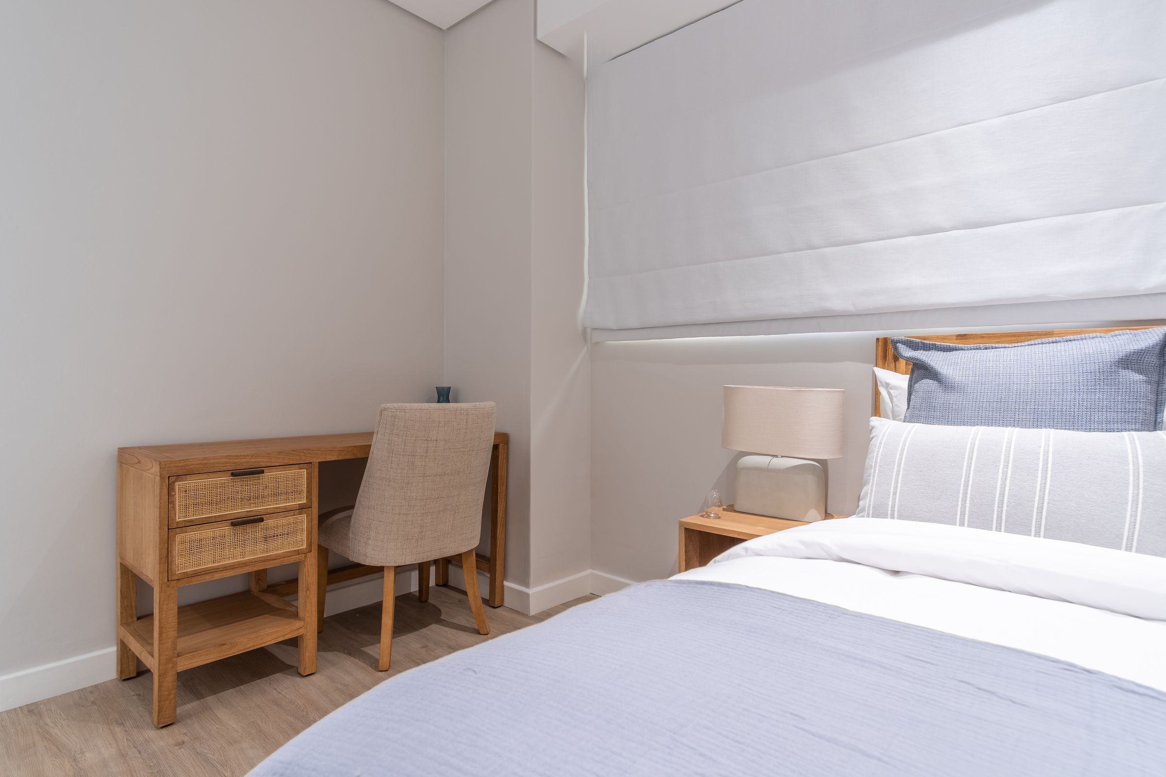 A cozy bedroom featuring a comfortable bed with light blue bedding, a bedside table with a lamp, and a wooden desk paired with a cushioned chair. The room is softly lit, with a large window covered by a white blind, creating a serene atmosphere.