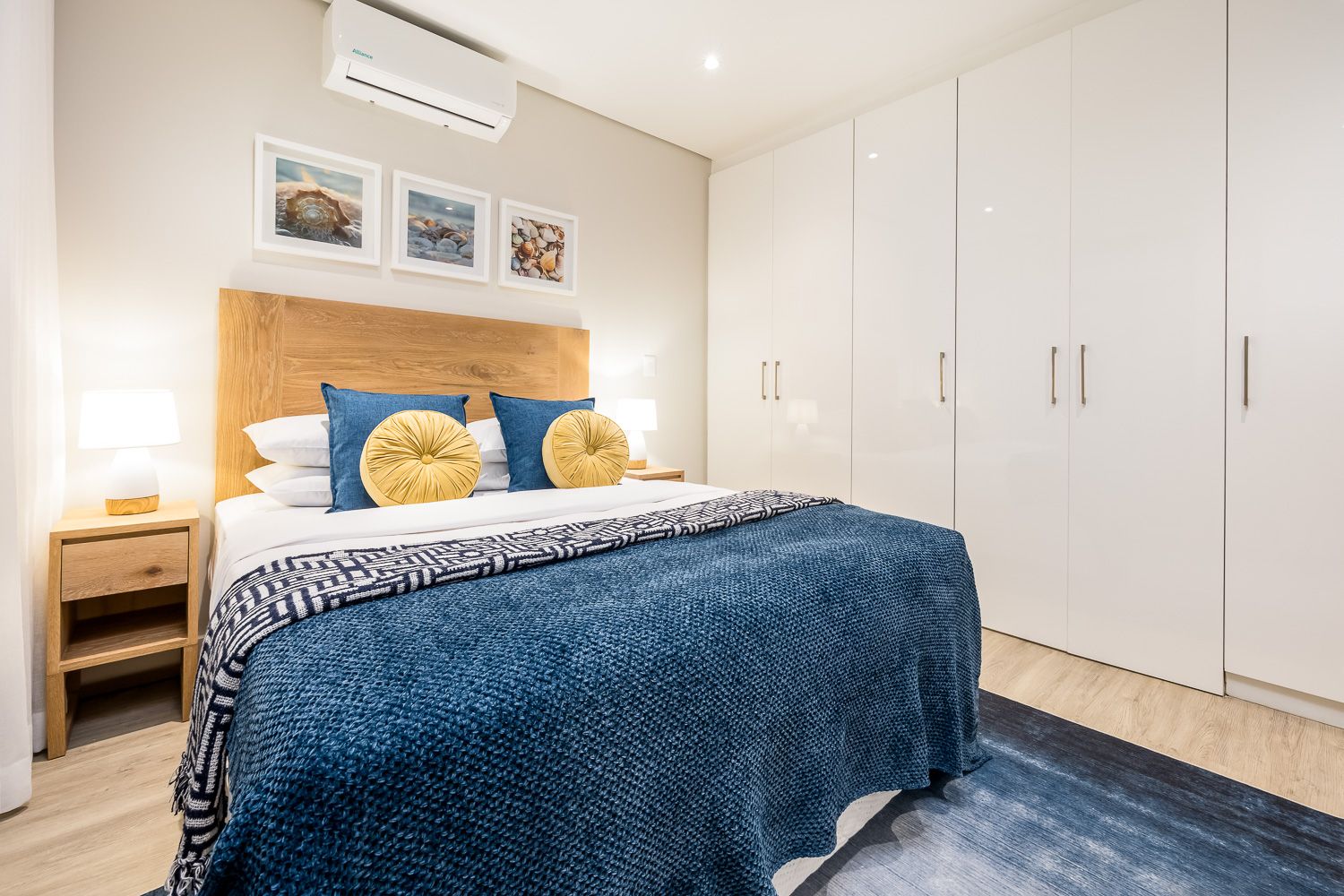 A cozy bedroom with a wooden headboard and blue bedding, accented by yellow cushions. White wardrobes line the wall, and soft lighting from bedside lamps creates a warm ambiance. Coastal artwork decorates the wall above the bed, enhancing the seaside theme.