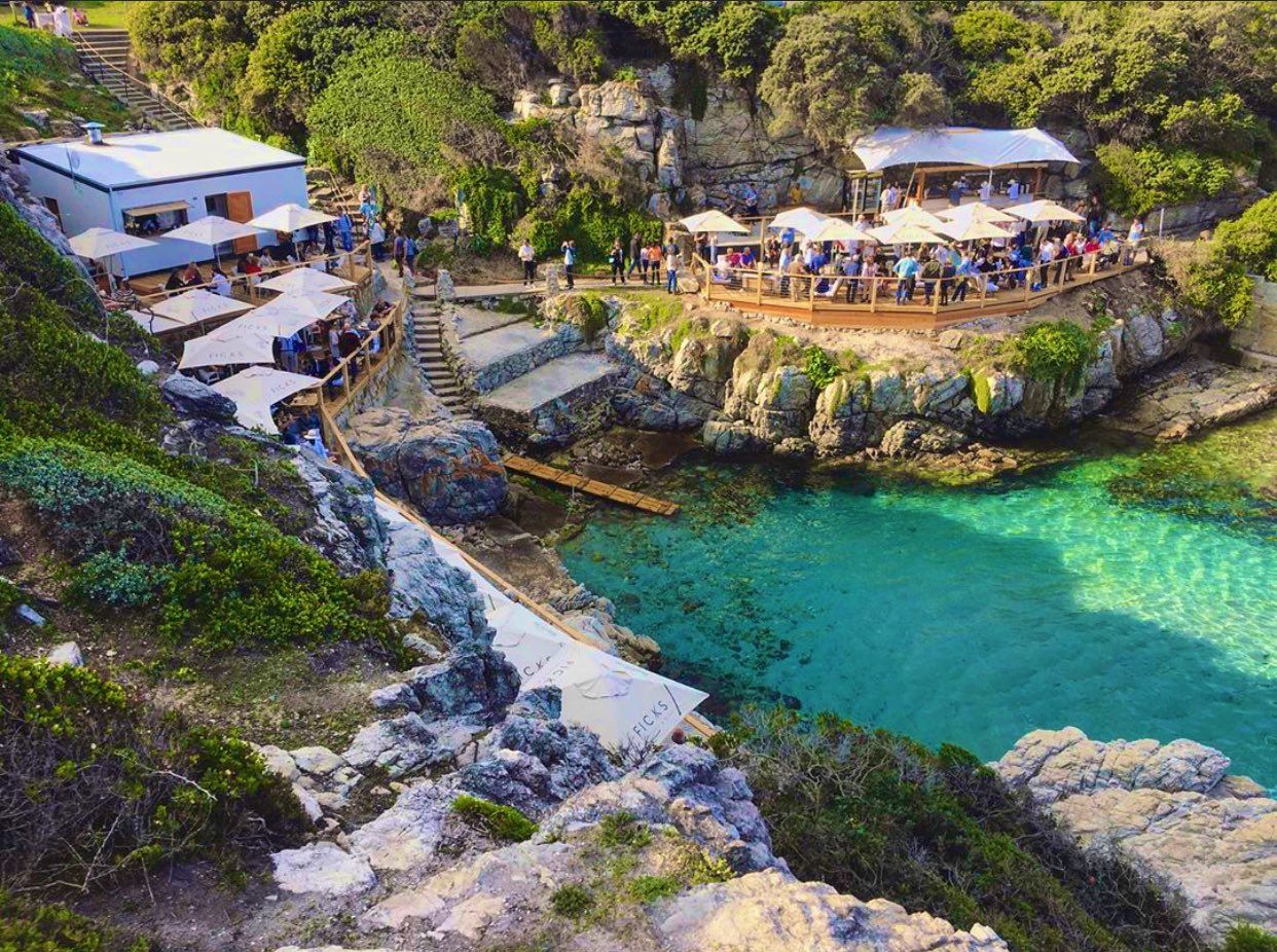 Aerial view of Ficks Pool, a cliffside tapas and cocktail bar in Hermanus, South Africa. The bar features outdoor seating with umbrellas, overlooking clear turquoise waters and rugged cliffs, creating a vibrant and scenic atmosphere for dining and socializing.