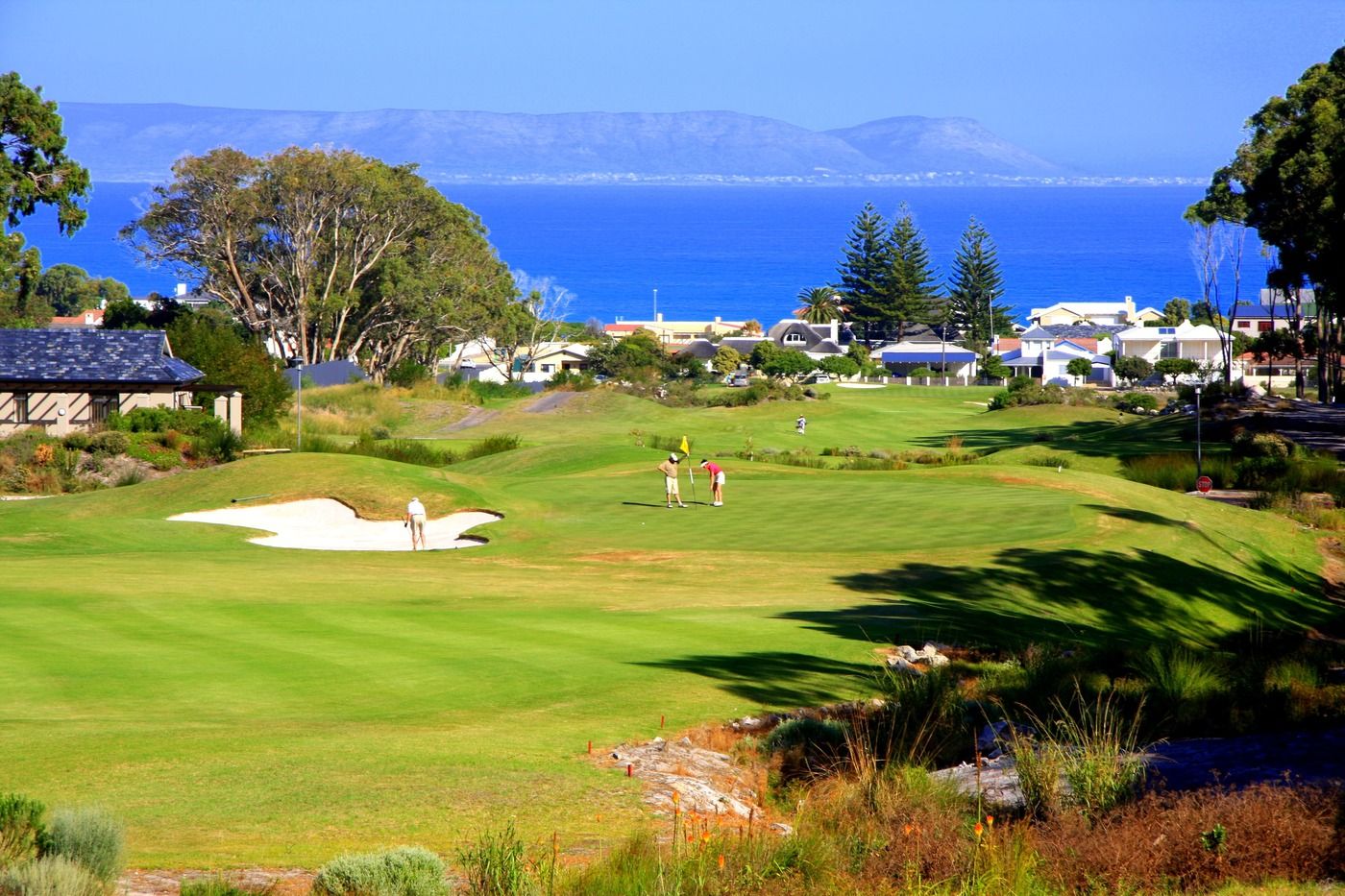 A vibrant golf course with players putting on the green, surrounded by lush greenery and sand bunkers. In the background, a coastal town and blue ocean are visible, with a mountain range in the distance under a clear blue sky.