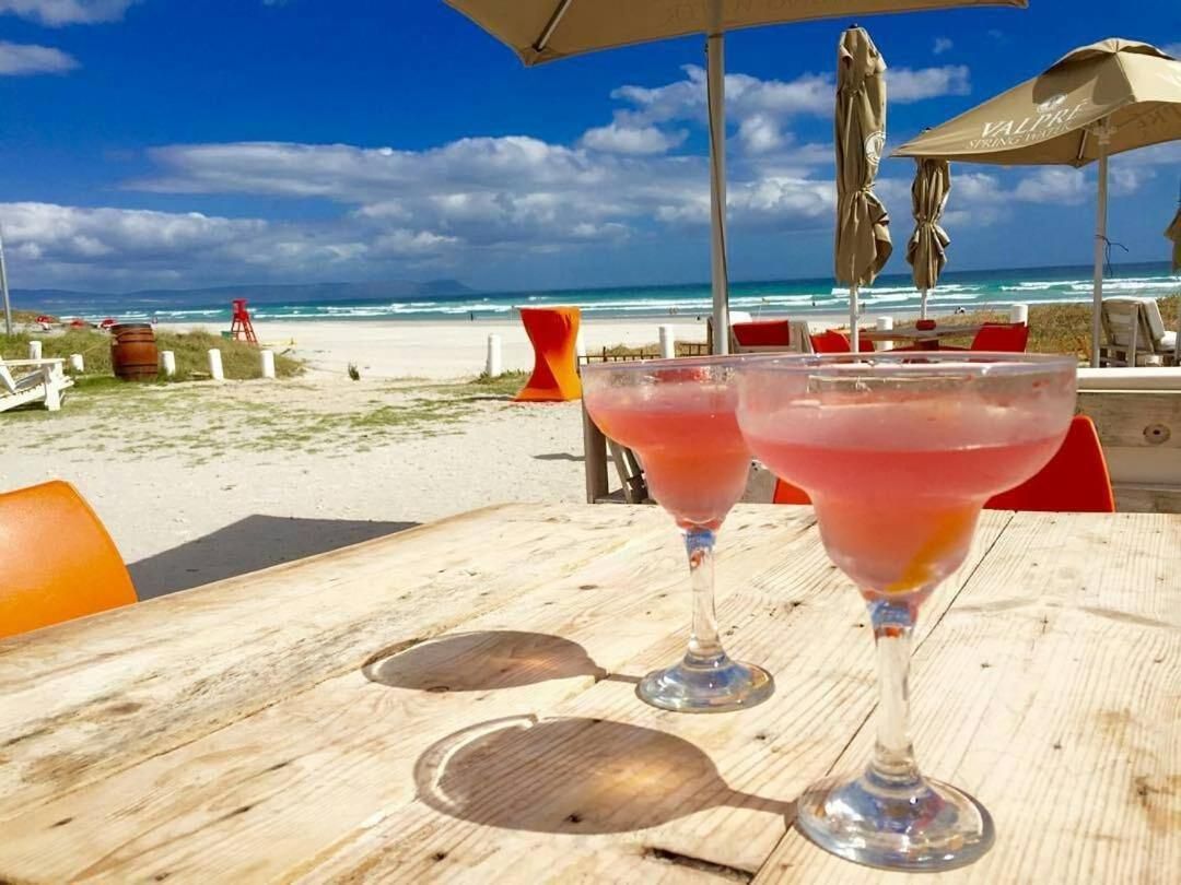 Two pink cocktails in glass goblets sit on a wooden table under umbrellas at a beachside location. The sandy beach and ocean waves are visible in the background, with a bright blue sky and scattered clouds overhead.