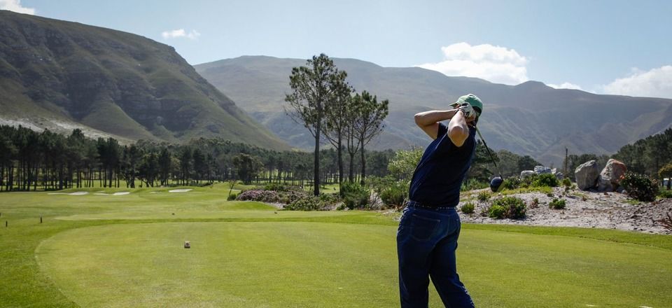 A golfer swings on a lush green course with a mountainous backdrop. The sky is clear with a few clouds, trees line the fairway, and the landscape is serene and picturesque.
