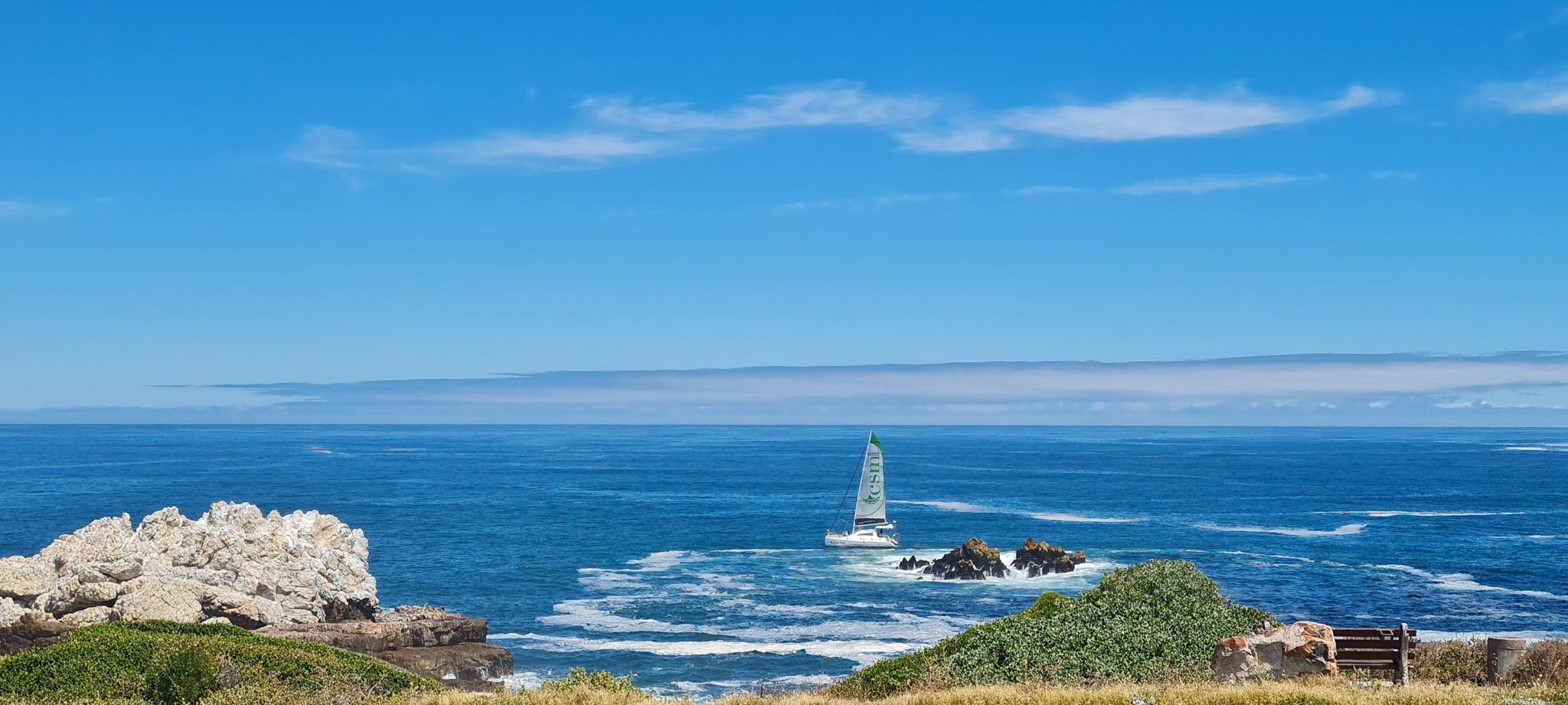 A sailboat with a green sail navigates the blue ocean near rocky formations. White clouds stretch across the clear sky, and a grassy area with a bench is visible in the foreground.