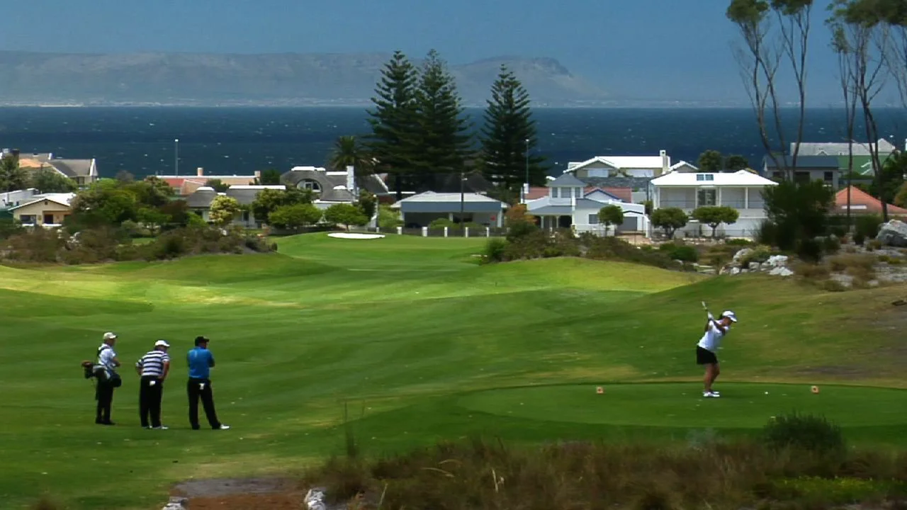 A golfer in mid-swing on a lush green golf course near the coast. Three people watch nearby, with houses and tall trees in the background. The ocean and distant mountains are visible under a clear blue sky.