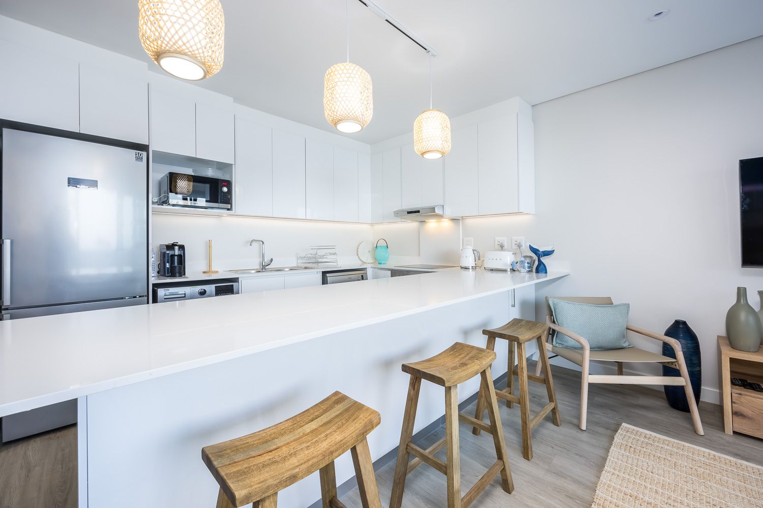 A sleek kitchen area featuring a long white countertop with wooden stools, pendant lighting, and modern appliances including a refrigerator, microwave, and coffee maker. The space is bright and airy, with minimalist decor and a cozy seating area with a cushion and decorative vases.