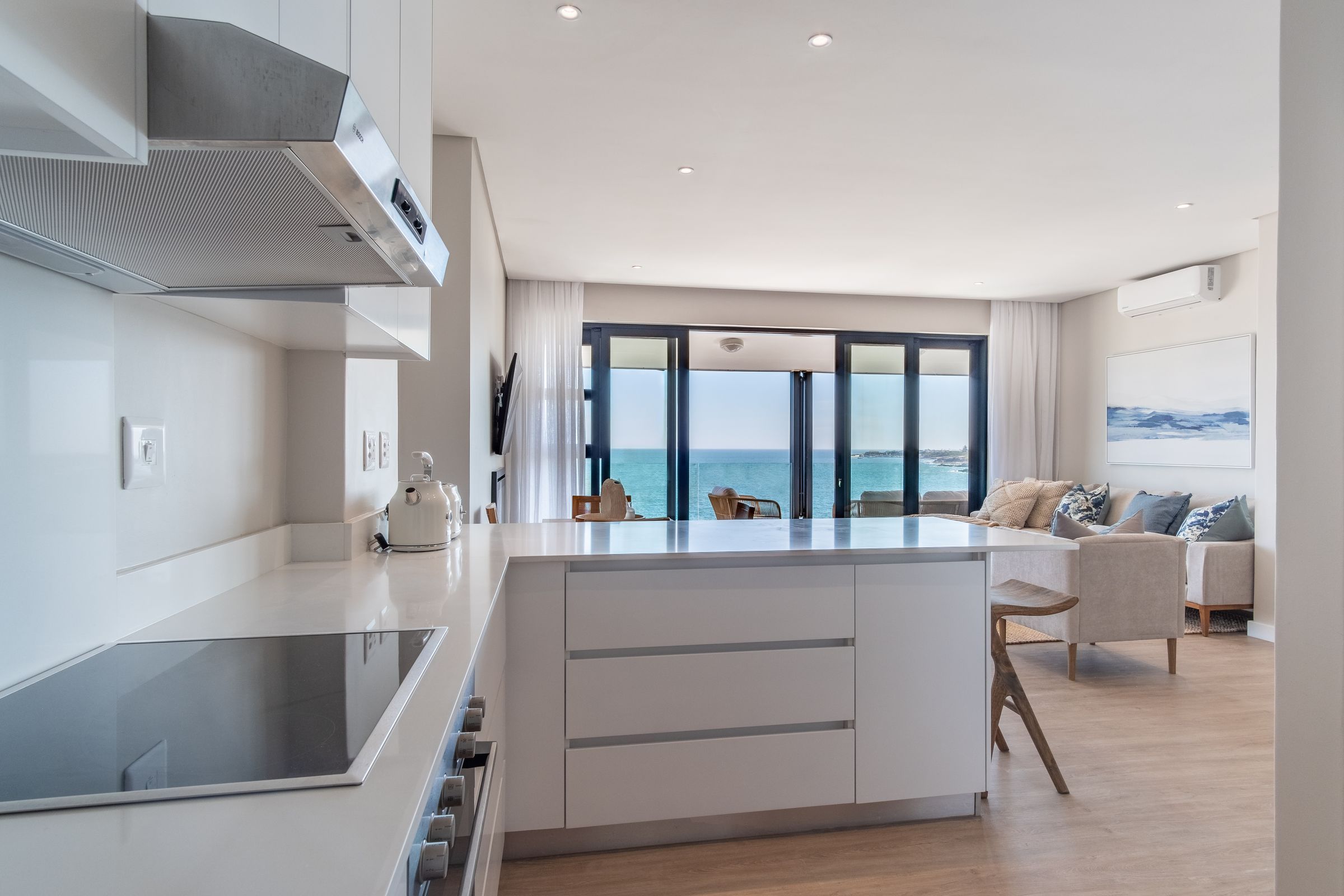 A sleek, modern kitchen with white cabinetry and a central island. The room opens into a living area with a large window offering a stunning view of the ocean. Light wood flooring and neutral-toned furniture create a serene atmosphere, complemented by coastal artwork on the wall.