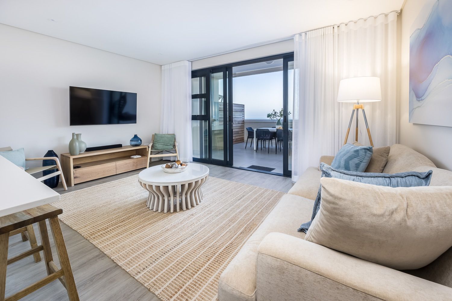A cozy living area featuring a beige sofa with blue cushions, a unique round coffee table, and a wall-mounted TV. The space is accented by a large woven rug, modern wooden furniture, and a floor lamp. Glass doors open to a dining area with a sea view, enhancing the coastal ambiance.
