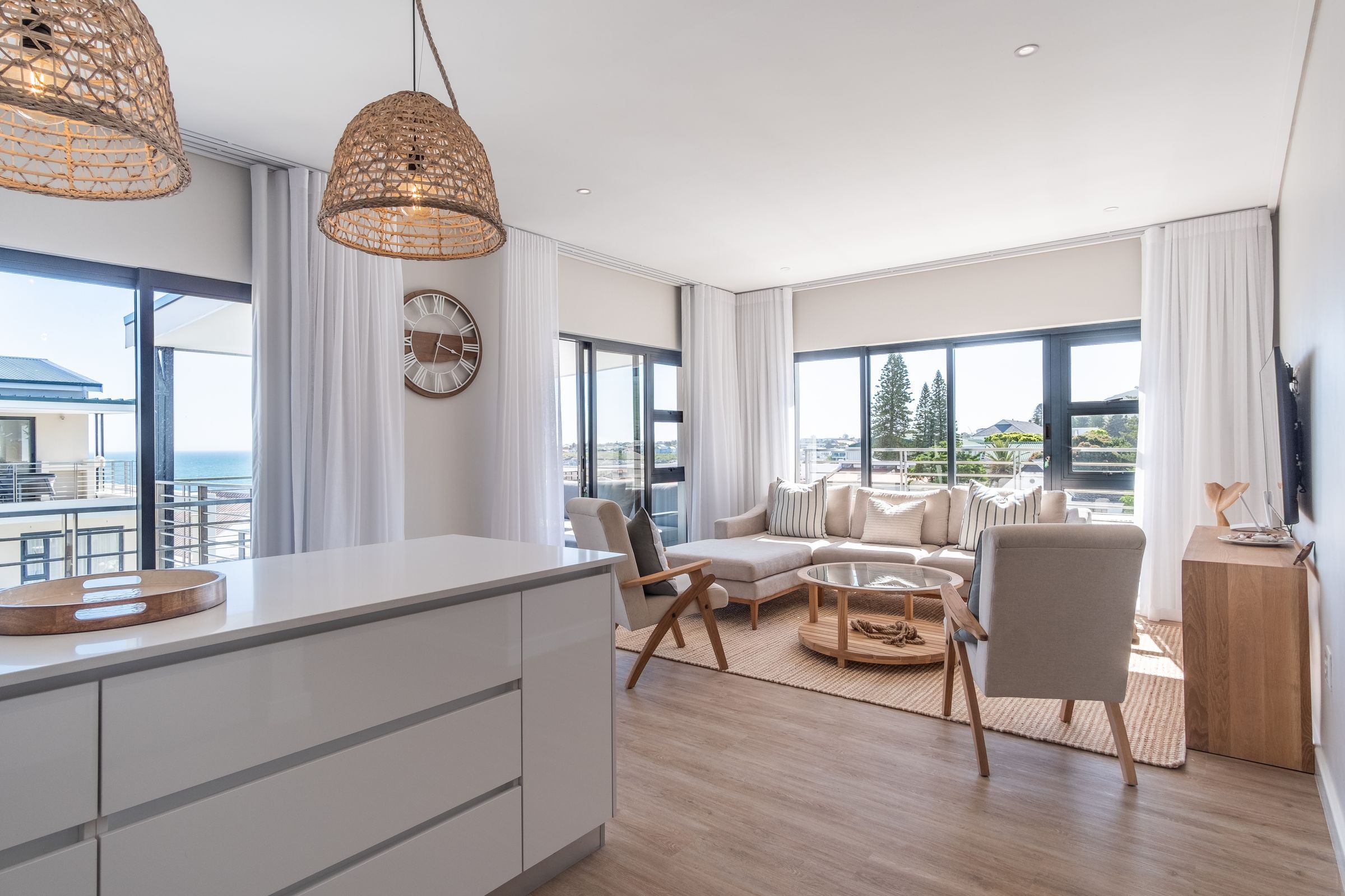 A modern living room featuring a beige sectional sofa, wooden chairs, and a glass coffee table. Large windows offer a partial sea view, with natural light filling the space. The decor includes wicker pendant lights and a wall-mounted TV, creating a cozy, inviting atmosphere.