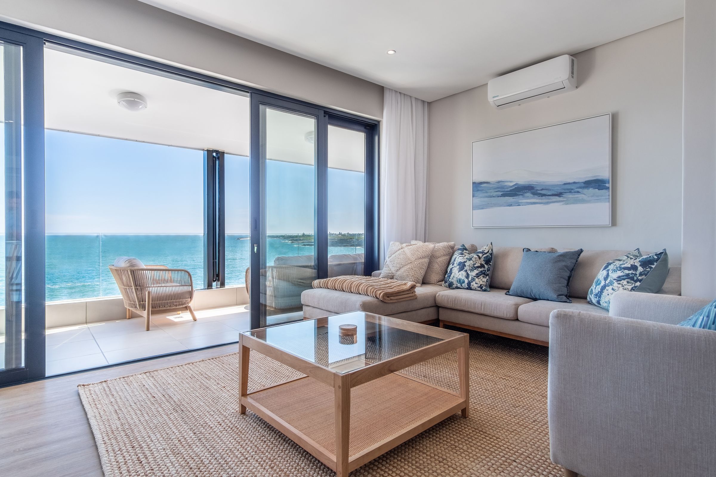 A spacious living room featuring a large L-shaped couch with blue and beige cushions, a glass-top coffee table, and a woven rug. Floor-to-ceiling sliding glass doors open to a balcony with stunning ocean views, enhancing the room's light and airy atmosphere.