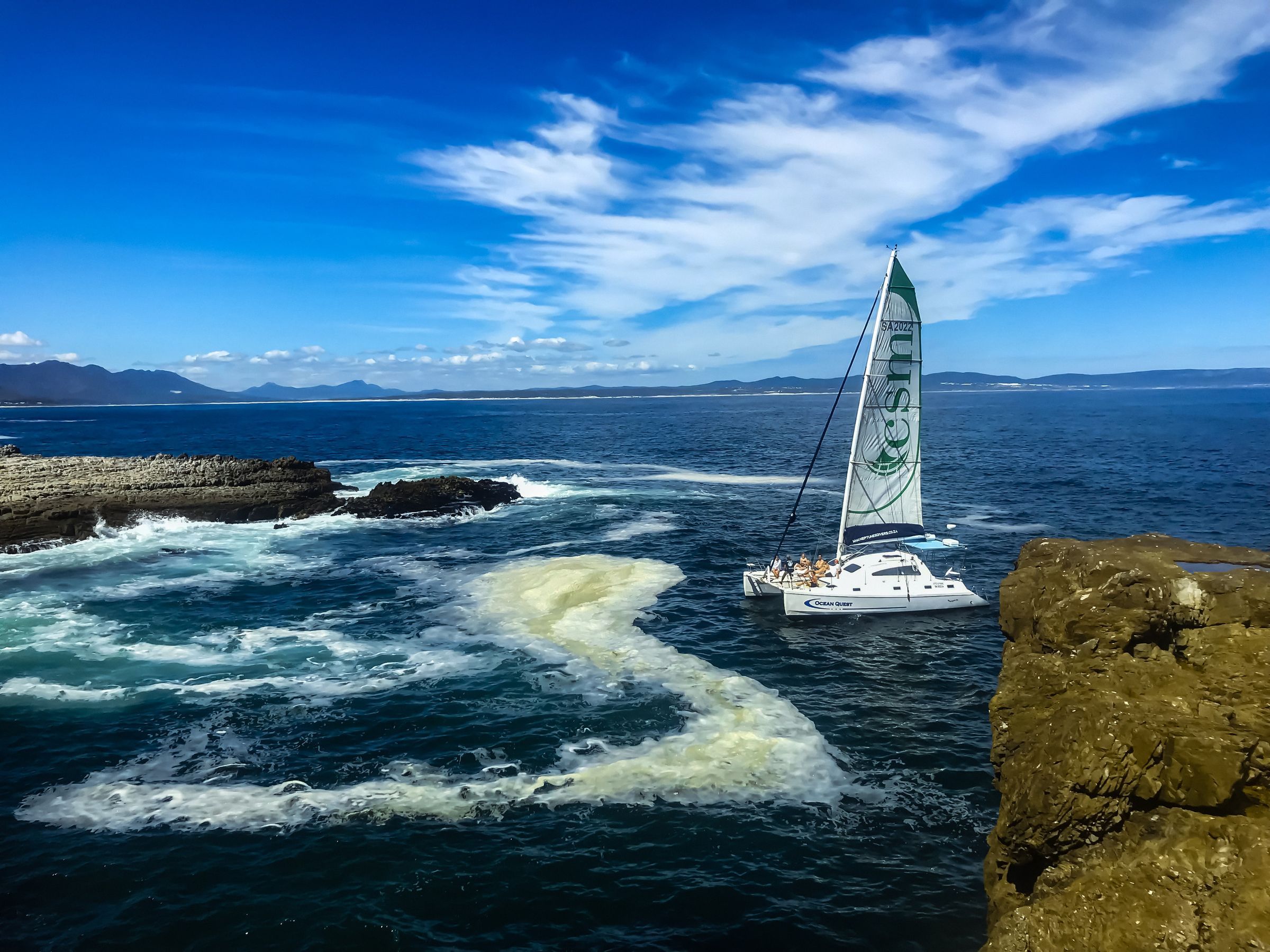 A sailboat with a tall, green sail navigates through turquoise waters near rocky cliffs. The sky is clear with wispy clouds, and distant mountains are visible on the horizon. The ocean waves create white foam against the dark rocks, enhancing the natural beauty of the scene.