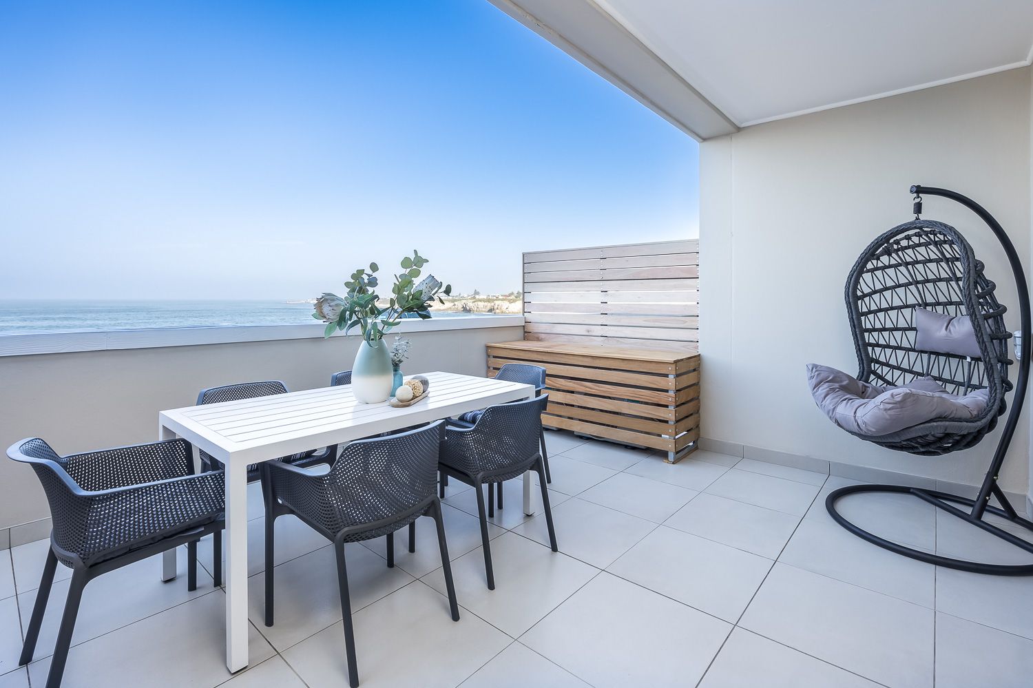 A modern balcony with ocean views features a white dining table set with black chairs, a hanging egg chair, and a decorative vase with greenery. The space is bright and airy, offering a relaxing coastal atmosphere.
