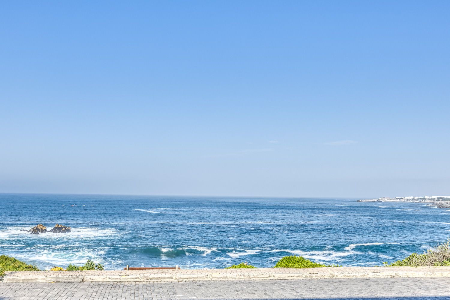 A scenic view of a vast blue ocean from a balcony, with gentle waves and a clear sky. The coastline stretches into the distance, bordered by greenery and a stone pathway in the foreground.