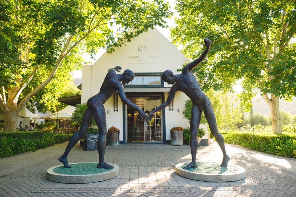 Two bronze sculptures depicting figures leaning towards each other, positioned on circular pedestals in front of a winery entrance. The scene is surrounded by lush green trees and a sunny pathway leading to the building.