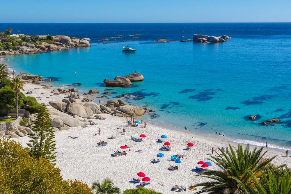 A stunning view of a Cape Town beach featuring pristine white sand, clear turquoise waters, and scattered boulders. Brightly colored umbrellas and sunbathers dot the shoreline, with lush greenery framing the scene. A boat floats peacefully in the azure sea under a clear blue sky.