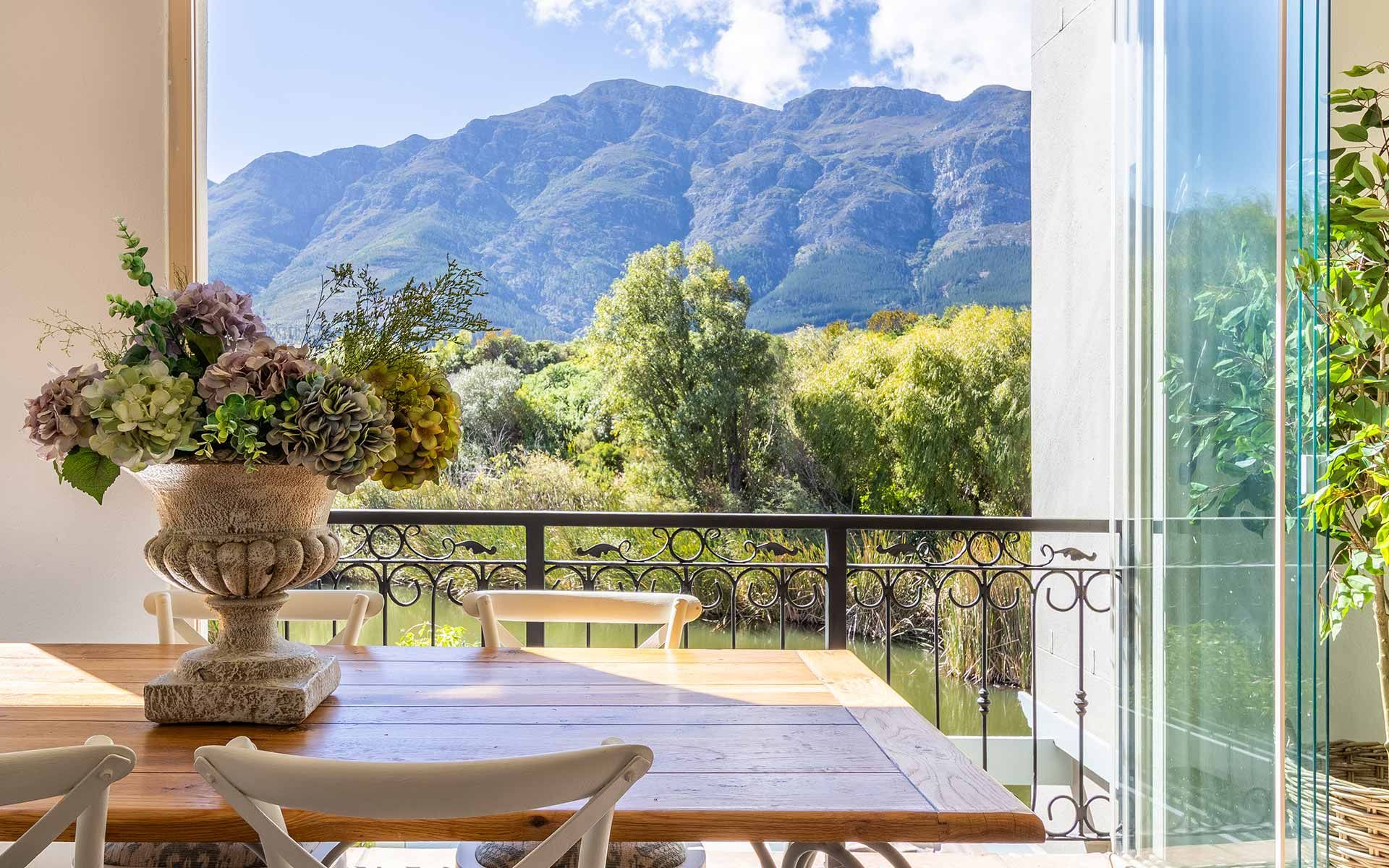 A wooden dining table with a stone vase of flowers overlooks lush greenery and a serene lake, framed by majestic mountains under a blue sky. The setting offers a tranquil and scenic view, perfect for enjoying meals in a peaceful environment.