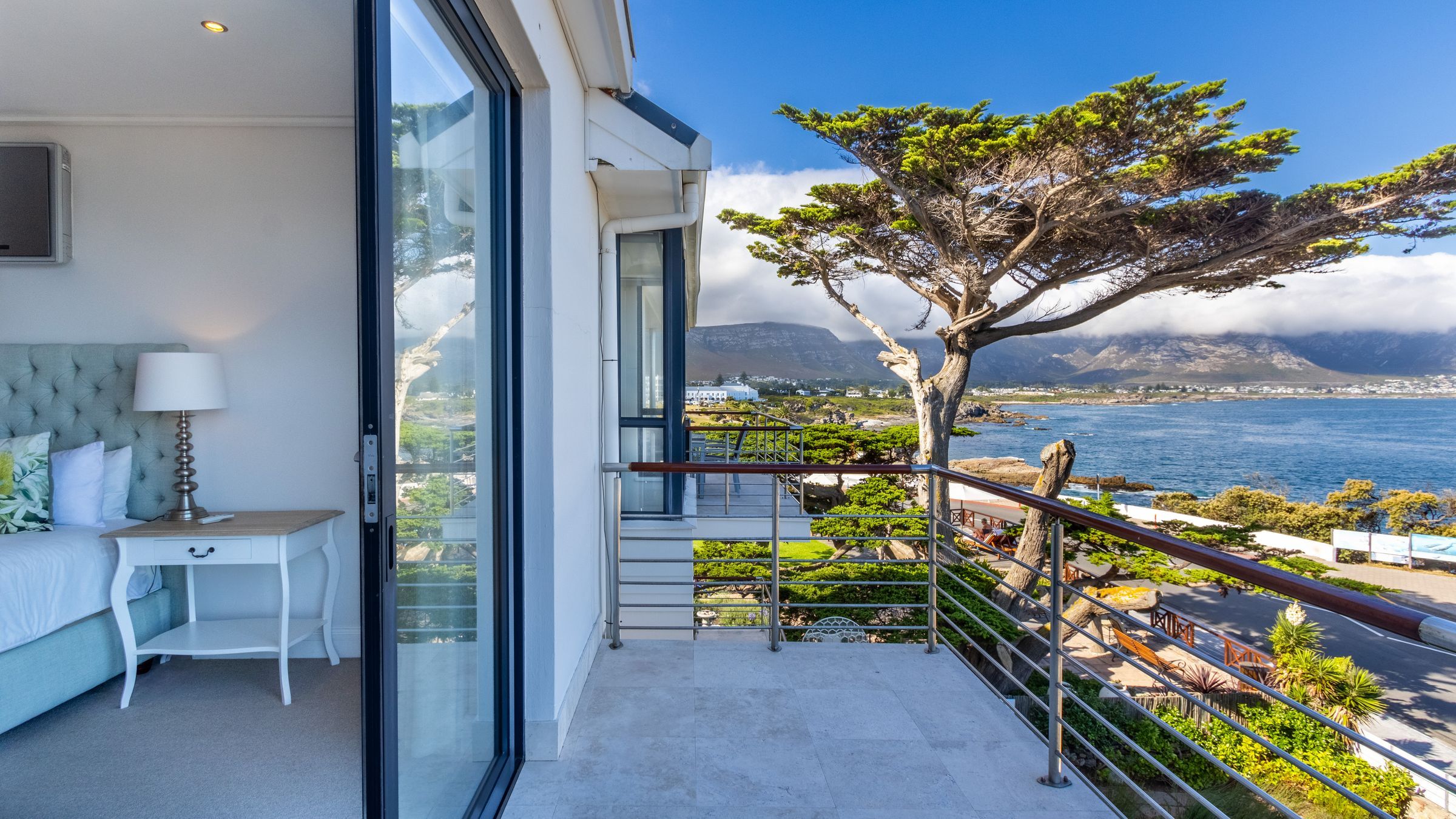 A modern bedroom with a large glass door opens to a balcony overlooking a scenic coastal view. The ocean, surrounded by greenery and distant mountains, is visible under a clear blue sky. A bed with pillows and a bedside table with a lamp are partially shown inside.
