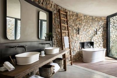 A bathroom featuring a rustic stone wall, twin white basins on a wooden counter, and a freestanding bathtub. Large mirrors and a wooden ladder with towels add to the decor. The room is well-lit by natural light from a large window, enhancing the earthy tones and textures.