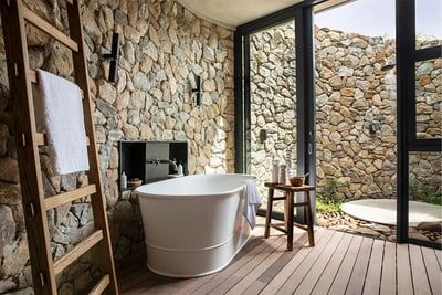 A cozy bathroom featuring a white freestanding tub against a textured stone wall. A wooden ladder holds a towel, and a small wooden stool sits nearby. Large windows offer a view of a stone patio, creating a natural, serene atmosphere.