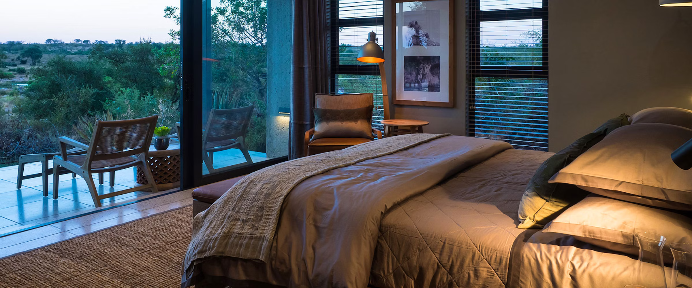 A stylish bedroom featuring a plush bed, wooden chairs, and a cozy reading nook. Large windows reveal a picturesque view of the lush landscape and distant horizon, offering a tranquil and inviting atmosphere.