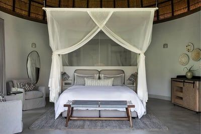 Stylish bedroom with a cozy double bed and white canopy. Features a round mirror, armchair, textured cushions, and a wooden bench. Decor includes woven baskets and a vase on a sideboard, creating a serene ambiance.