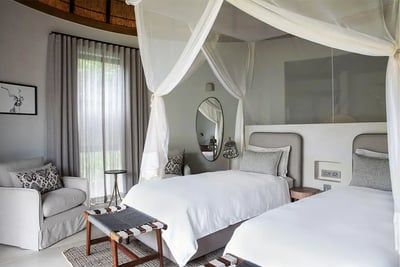 A stylish bedroom with two twin beds featuring white linens and a sheer canopy. The room includes a cushioned chair, a small round table, and a wall mirror. A window with sheer curtains allows natural light, and the decor includes a monochrome wall art piece.