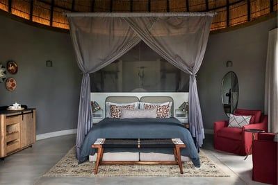 A stylish bedroom with a canopy bed draped in sheer gray fabric. The bed has patterned pillows and a dark blue comforter. A red armchair sits to the right, with a round mirror above a small table. Decor includes wall art and a rug, creating a cozy, modern ambiance.