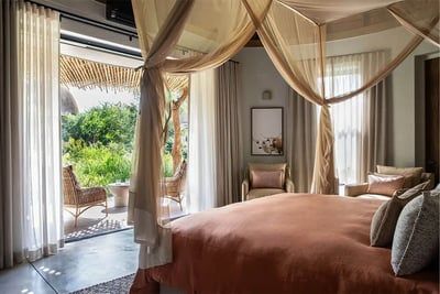 A luxurious bedroom featuring a canopy bed with beige curtains, soft bedding, and plush pillows. The room opens to a balcony with wicker chairs and a view of lush greenery, allowing natural light to stream in through sheer curtains. A serene and inviting atmosphere is created.